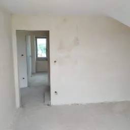 Surowe ściany po nałożeniu tynku w nowym, jasnym pomieszczeniu z widocznymi otworami pod gniazdka elektryczne i kable zwisające ze ściany, widok na kolejne pomieszczenie przez otwarte drzwi.