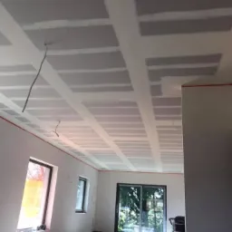 Sufit z widoczną konstrukcją z profili i płyt gipsowo-kartonowych przed szpachlowaniem spoin, w nowym pomieszczeniu z dużym oknem balkonowym i dwoma mniejszymi oknami, z pomarańczową linią...