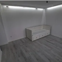 Pomalowane na biało wnętrze pokoju z szarą podłogą imitującą drewno i białą sofą z trzema szufladami, oświetlone listwami LED pod sufitem.