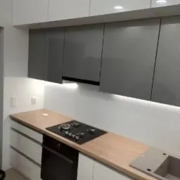 Nowoczesna kuchnia: białe szafki z szarymi frontami, drewniany blat, płyta gazowa i zlewozmywak w minimalistycznym stylu.