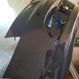 Pomalowany na ciemny szary kolor zderzak samochodowy, widoczne refleksy światła, w tle folia malarska i fragment ściany.