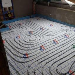 Hydro-gaz Krystian Ostrów WLKp - Instalacja ogrzewania podłogowego w poddaszu: białe rury grzewcze ułożone spiralnie na niebieskiej siatce, widoczny fragment okna dachowego i niezagospodarowana przestrzeń pod skosem.