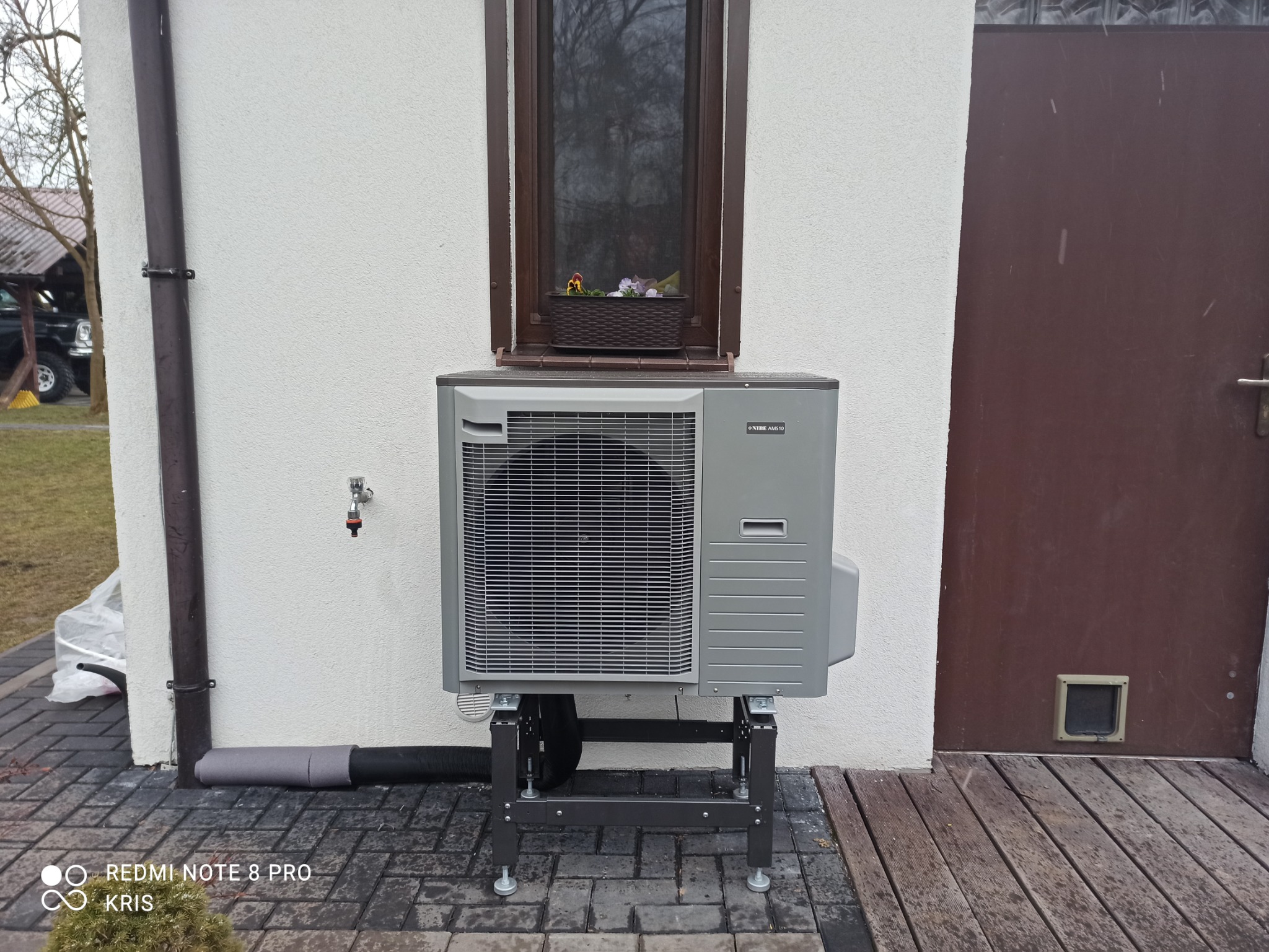 Szara pompa ciepła NIBE AMS10 zamontowana na metalowym stelażu przy białej ścianie budynku z brązowymi drzwiami i oknem z kwiatami.