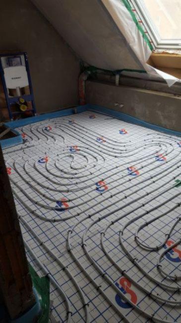 Instalacja ogrzewania podłogowego w poddaszu: białe rury grzewcze ułożone spiralnie na niebieskiej siatce, widoczny fragment okna dachowego i niezagospodarowana przestrzeń pod skosem.