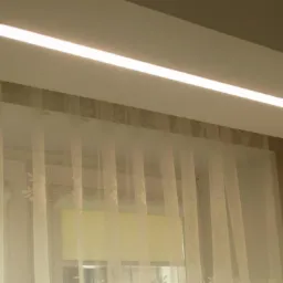 Nowoczesna, długa lampa LED wpuszczona w sufit, oświetlająca pomieszczenie z oknem zasłoniętym przez delikatne, białe firanki z motywem kwiatowym.