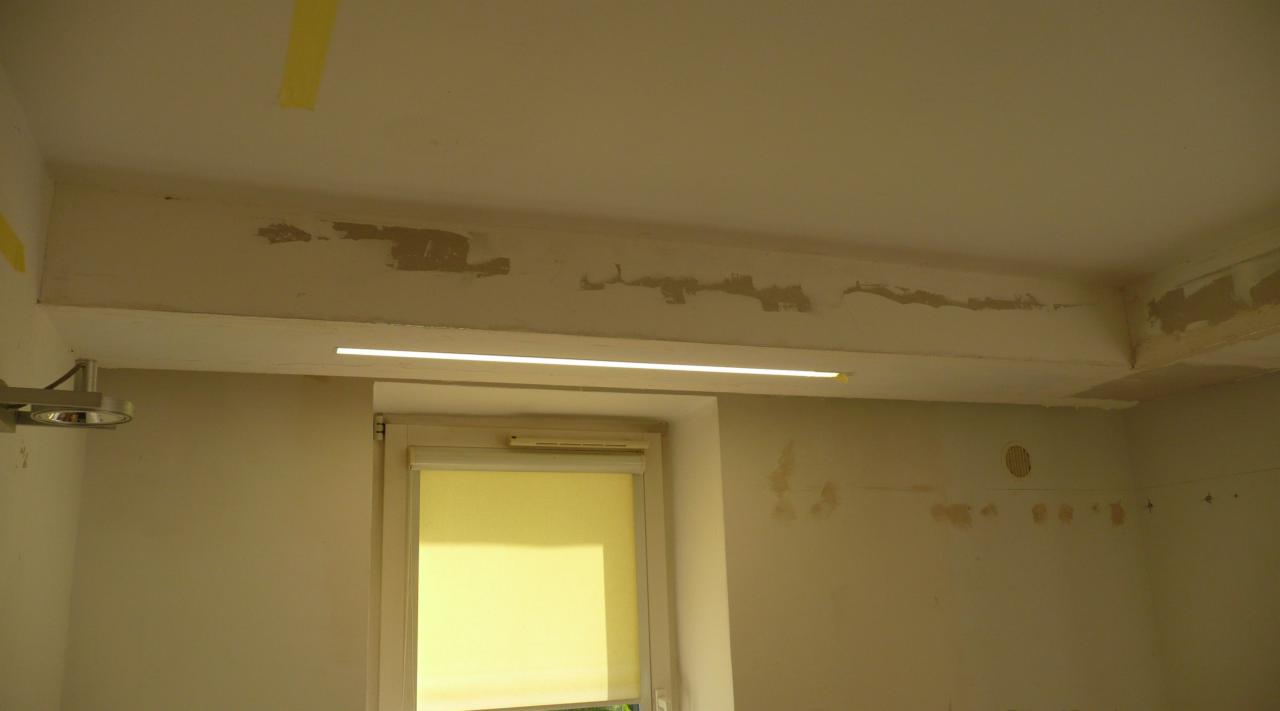 Wnętrze pokoju w trakcie remontu, widoczna listwa LED pod sufitem, ślady po szpachlowaniu na ścianach i suficie, okno z roletą.