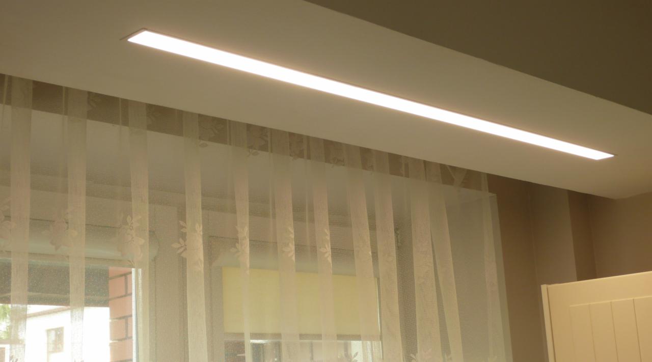 Nowoczesna, długa lampa LED wpuszczona w sufit, oświetlająca pomieszczenie z oknem zasłoniętym przez delikatne, białe firanki z motywem kwiatowym.