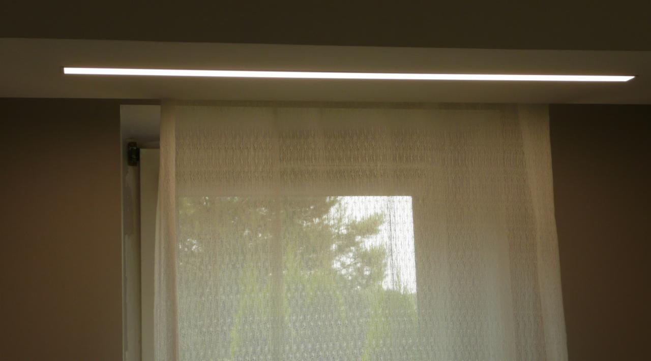Nowoczesna, długa lampa LED zamontowana pod sufitem, oświetlająca okno zasłonięte przez delikatną, kremową firankę. Widok na zieleń za oknem.