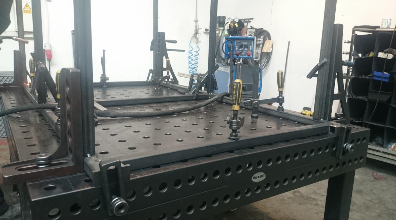 Spawanie ramy z profili stalowych na stole spawalniczym Siegmund, widoczne narzędzia i uchwyty mocujące, spawarka oraz osprzęt w tle.