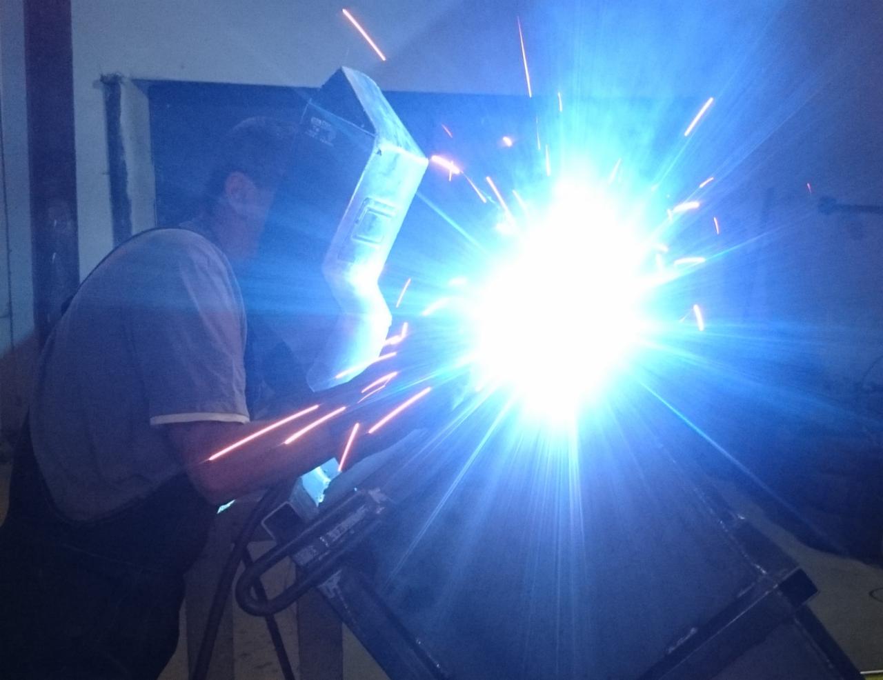 Pracownik w masce spawalniczej łączy elementy metalowe, iskry rozświetlają ciemne pomieszczenie warsztatowe.