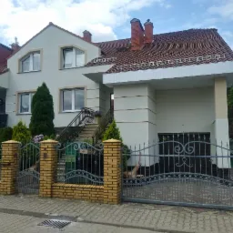 Dwupiętrowy dom z jasną elewacją, czerwonym dachem i bramą z ceglanymi słupkami, sfotografowany w pochmurny dzień.