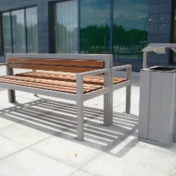 Nowoczesna ławka parkowa z drewnianym siedziskiem i metalową ramą, stojąca obok metalowego kosza na śmieci w szarym kolorze, na tle budynku z dużymi oknami.