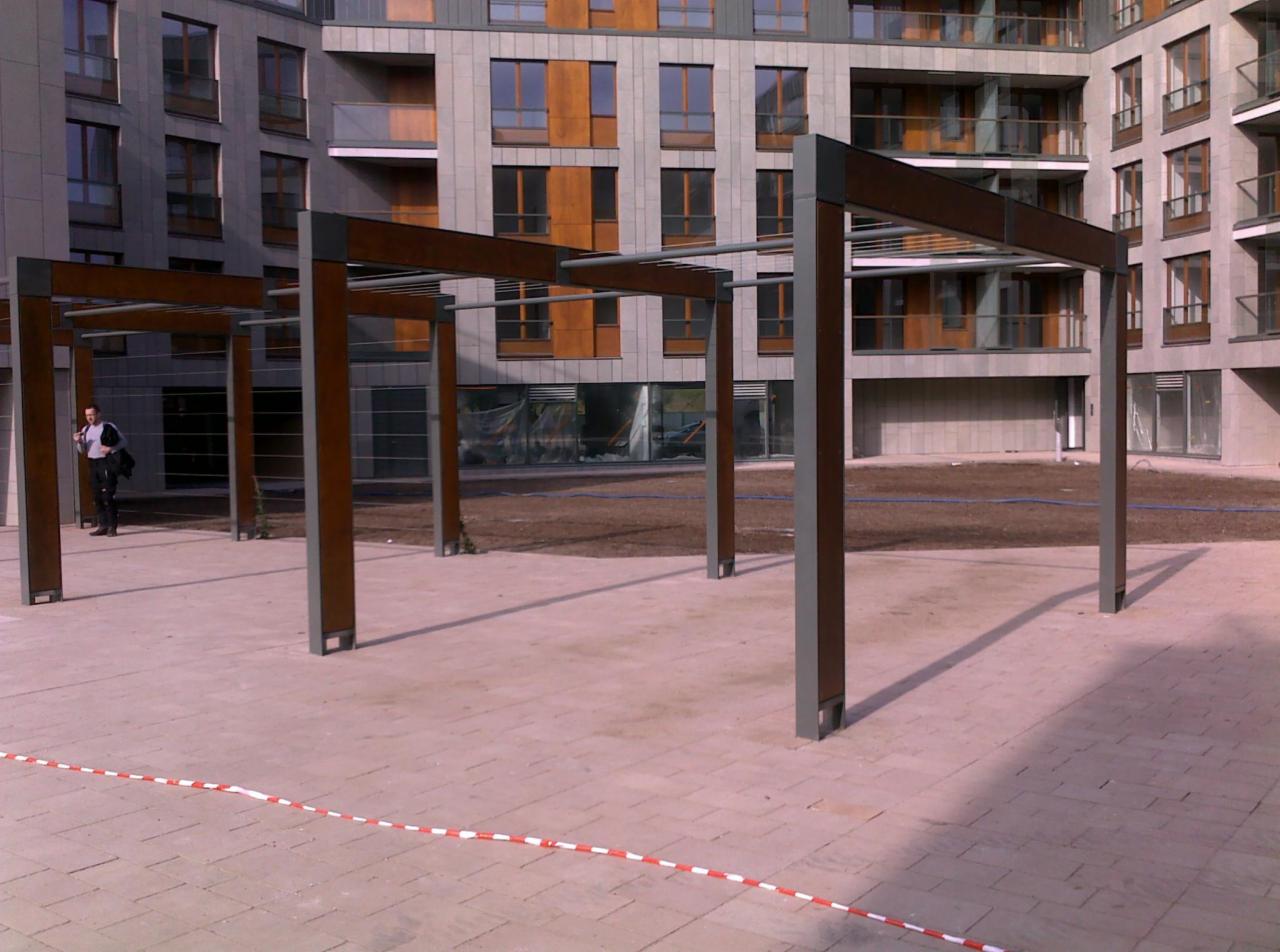 Metalowa pergola z drewnianymi elementami na placu przed budynkiem mieszkalnym, widoczny mężczyzna w tle i taśma ostrzegawcza.