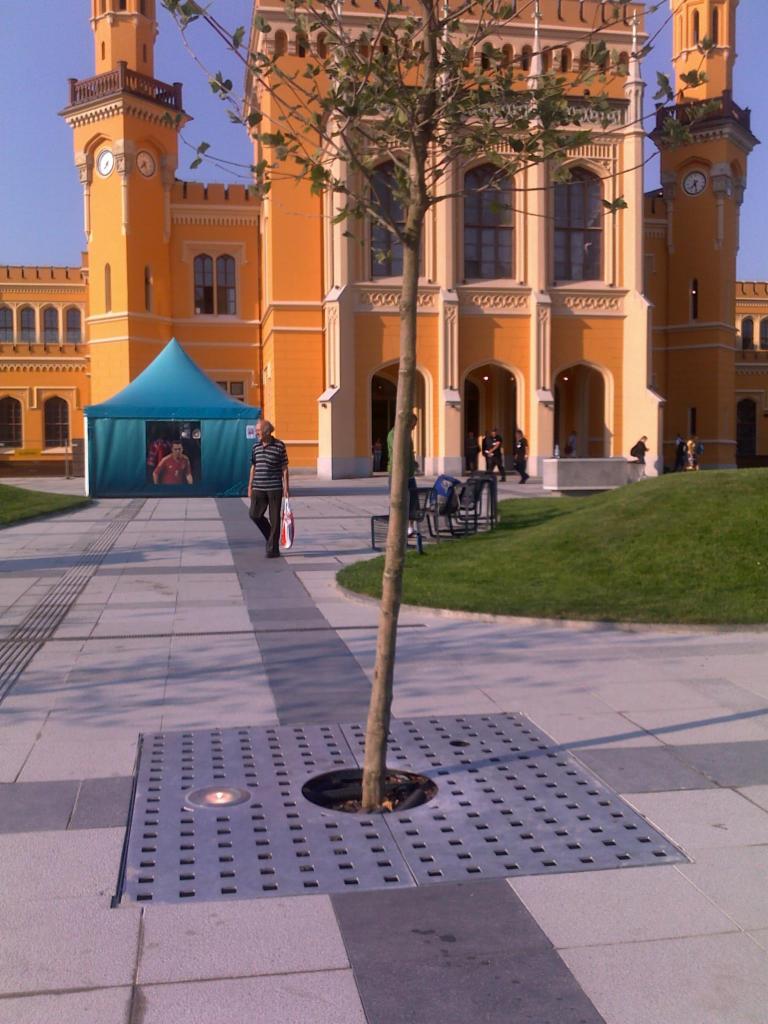 Żółty budynek usługowy z wieżami i zegarami, widoczny z poziomu chodnika z drzewem w metalowej osłonie i niebieskim namiotem z grafiką.