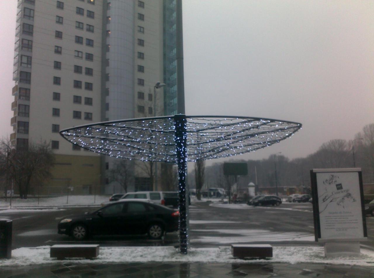 Metalowa konstrukcja w kształcie parasola, oświetlona białymi lampkami, na tle miejskiego krajobrazu w zimowej scenerii.