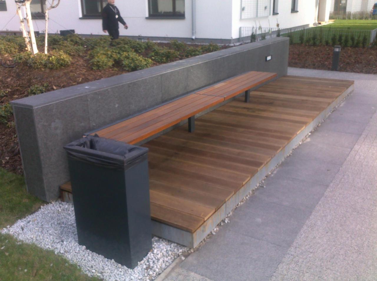 Zewnętrzna strefa relaksu z drewnianym podestem, ławką z metalowymi nogami i betonowym murem oporowym, uzupełniona o kosz na śmieci i oświetlenie, z widoczną osobą w tle.