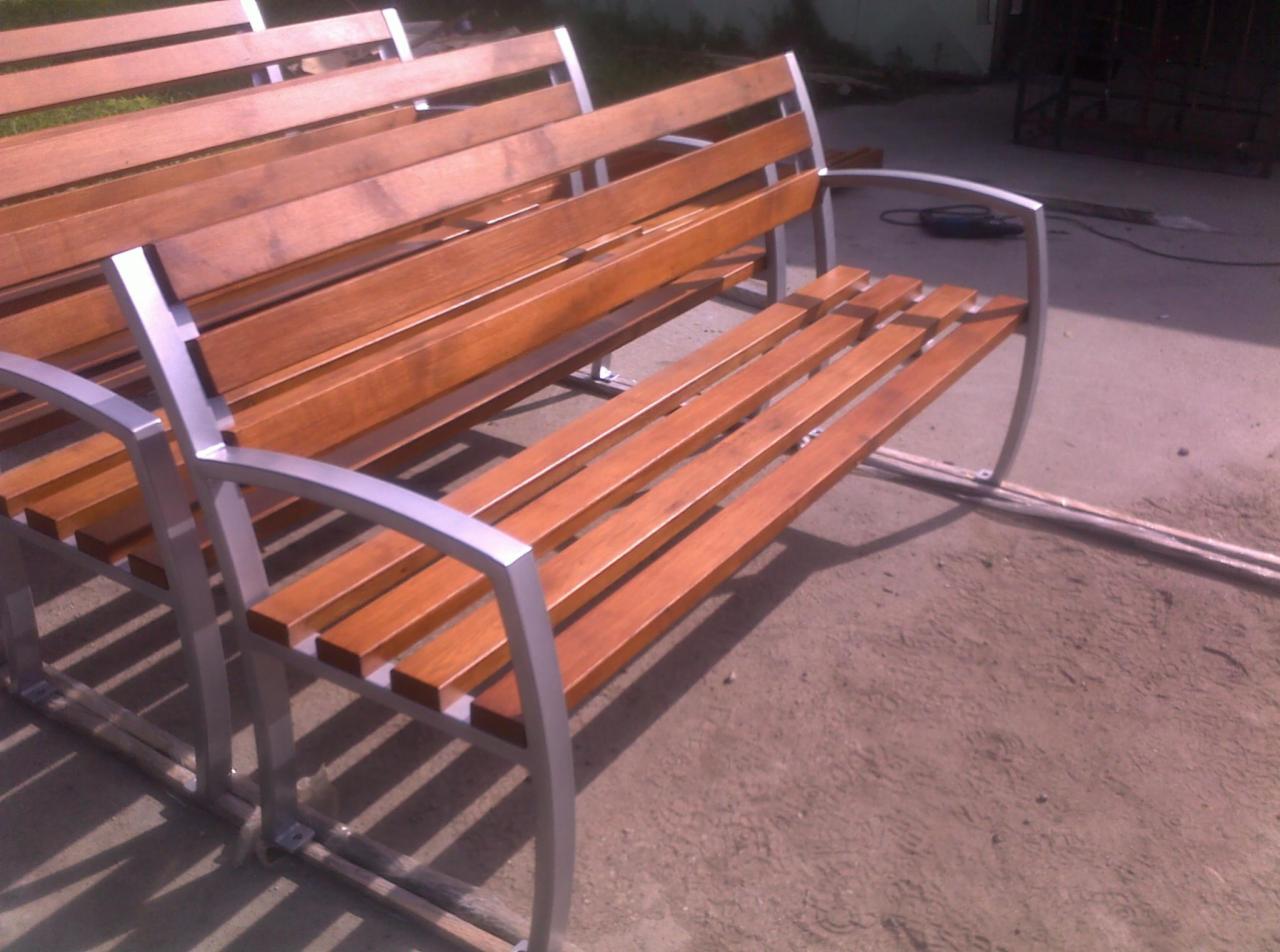 Dwie ławki parkowe z drewnianymi siedziskami i metalową, jasną ramą, ustawione w warsztacie lub na zewnątrz, widoczne ślady prac montażowych.