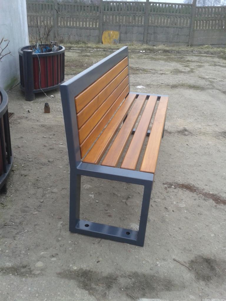 Ławka parkowa z drewnianym siedziskiem i oparciem na metalowym stelażu, ustawiona na betonowym podłożu obok donic z roślinami, w tle betonowe ogrodzenie.