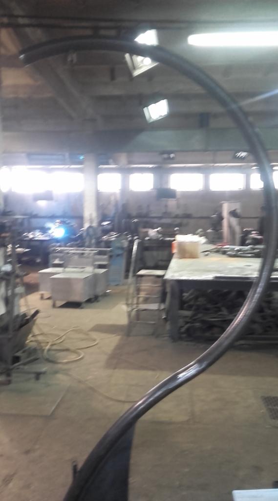 Wnętrze warsztatu z widocznym fragmentem wygiętej, metalowej rury, prawdopodobnie elementu konstrukcji stalowej, z pracującym spawaczem w tle.