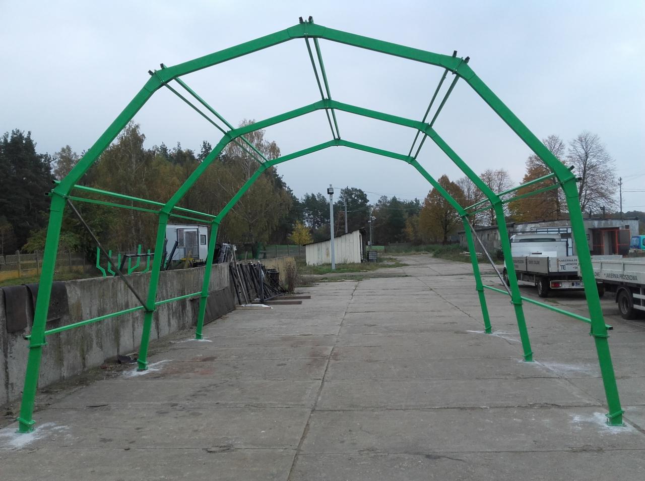 Zielona, łukowa konstrukcja stalowa w trakcie montażu na betonowej powierzchni, widoczne ślady po spawaniu u podstawy, w tle budynki i drzewa.