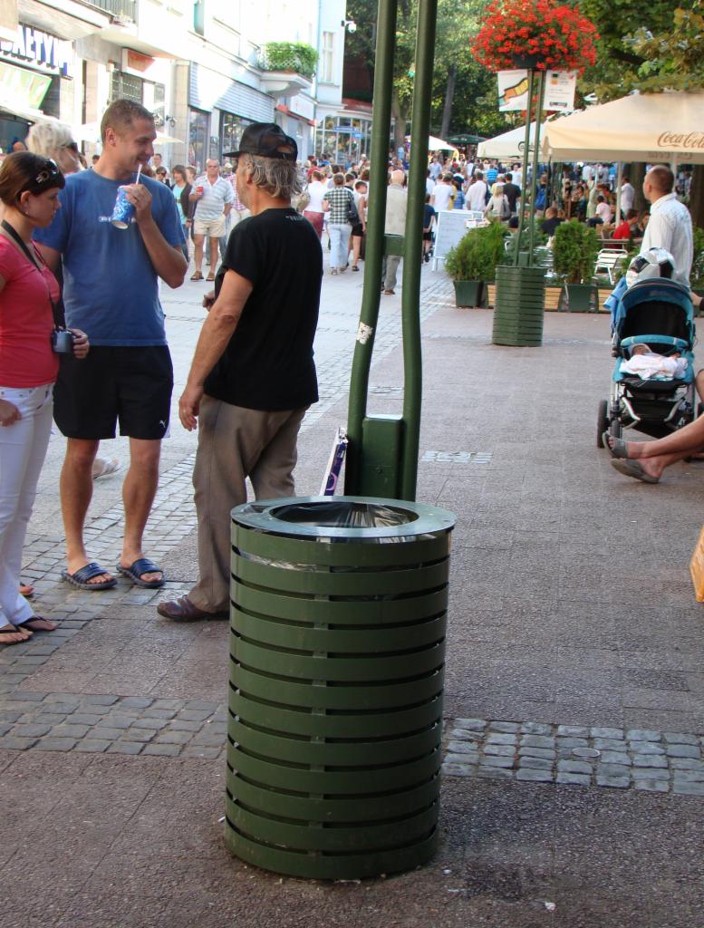 Zielony, metalowy kosz na śmieci na chodniku w miejskiej przestrzeni publicznej, otoczony przez przechodniów i elementy małej architektury.