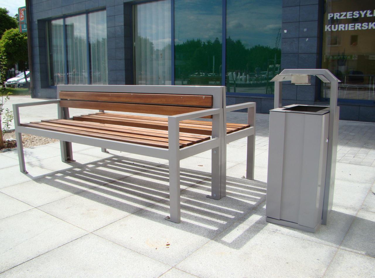 Nowoczesna ławka parkowa z drewnianym siedziskiem i metalową ramą, stojąca obok metalowego kosza na śmieci w szarym kolorze, na tle budynku z dużymi oknami.