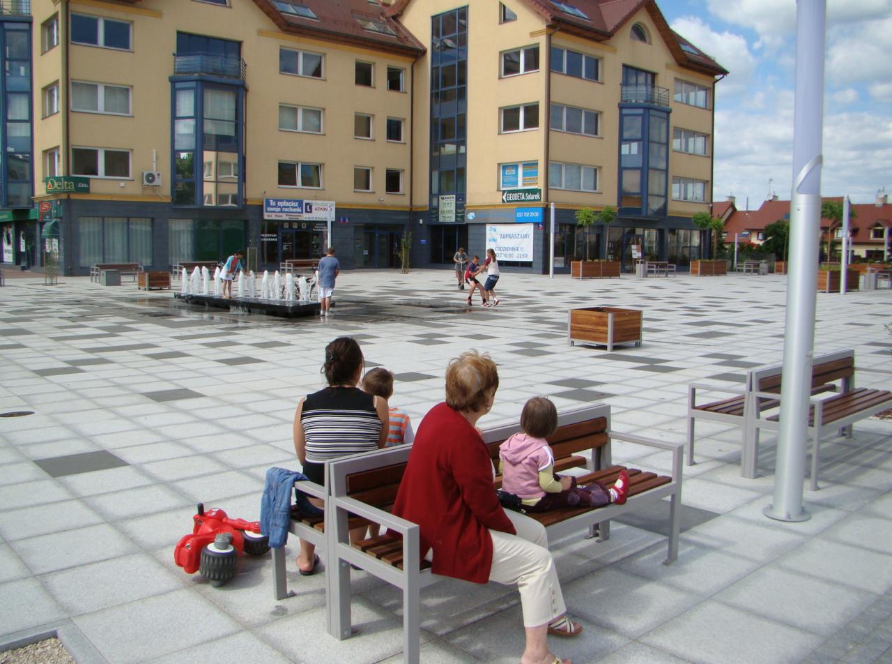 Miejski plac z fontanną i budynkami usługowymi w tle, na pierwszym planie ławki z odpoczywającymi osobami i czerwony rowerek biegowy.