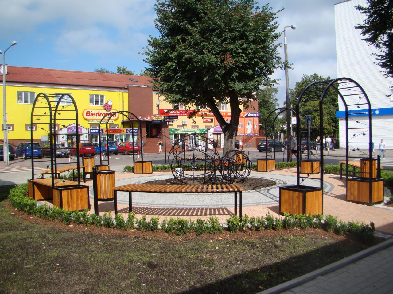Miejska przestrzeń rekreacyjna z ławkami, metalowymi konstrukcjami i kulistymi rzeźbami, otoczona niską zielenią na tle budynków handlowych.