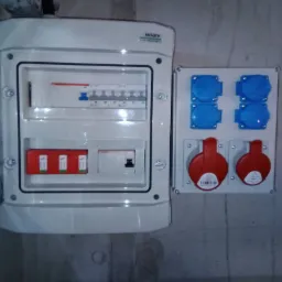 Rozdzielnica elektryczna z otwartymi drzwiczkami obok gniazd siłowych, zamontowana na szarej, surowej ścianie.