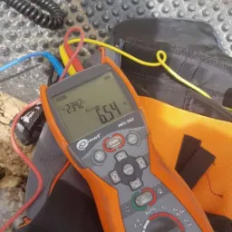 Miernik elektryczny Sonel MPI-502 z podłączonymi przewodami pomiarowymi, wyświetlający wynik pomiaru, leżący na torbie narzędziowej na tle teksturowanej powierzchni.