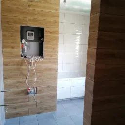 Wnętrze łazienki w trakcie remontu: ściany wyłożone płytkami imitującymi drewno, widoczny otwór na bojler z wystającymi przewodami, białe płytki na ścianie w głębi, szare płytki podłogowe, surowy...