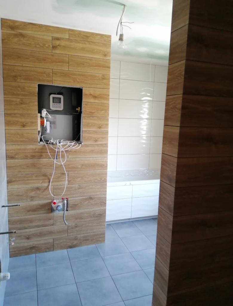 Wnętrze łazienki w trakcie remontu: ściany wyłożone płytkami imitującymi drewno, widoczny otwór na bojler z wystającymi przewodami, białe płytki na ścianie w głębi, szare płytki podłogowe, surowy...