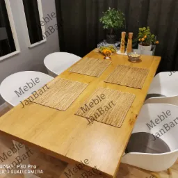 Stół z jasnego drewna z czterema plecionymi podkładkami, otoczony białymi krzesłami z szarymi poduszkami; w tle rośliny doniczkowe i minimalistyczne lustra.