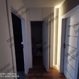 Wnętrze domu z trzema białymi drzwiami, oświetleniem LED wzdłuż ściany i widocznymi szafkami wiszącymi nad jednym z przejść.