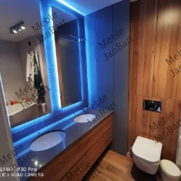 Nowoczesna łazienka z podświetlanym lustrem, drewnianymi szafkami pod umywalką z szarą powierzchnią, drewnianą ścianą i wiszącą toaletą.