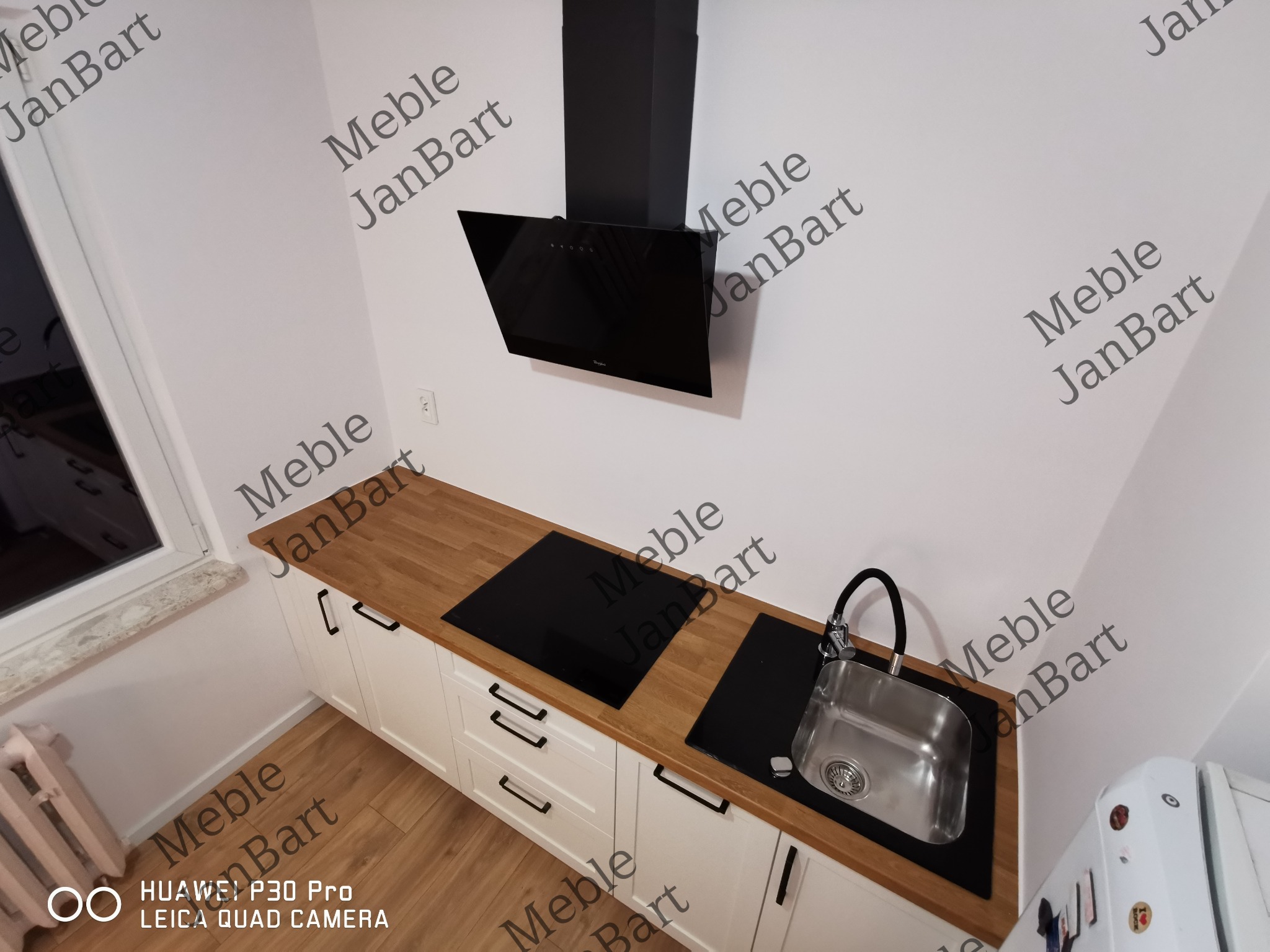 Nowoczesna zabudowa kuchenna z drewnianym blatem, czarną płytą grzewczą i zlewem ze stali nierdzewnej, białe fronty szafek z czarnymi uchwytami, czarny okap nad płytą, widoczny fragment lodówki.