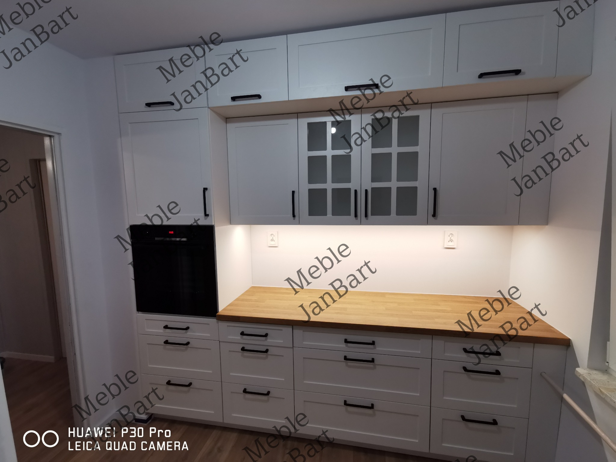 Biała zabudowa kuchenna z drewnianym blatem, czarnymi uchwytami i piekarnikiem w zabudowie, widok z dołu.