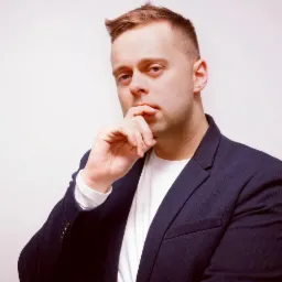 Portret mężczyzny w marynarce, zamyślony, z ręką przy ustach, na jasnym tle.