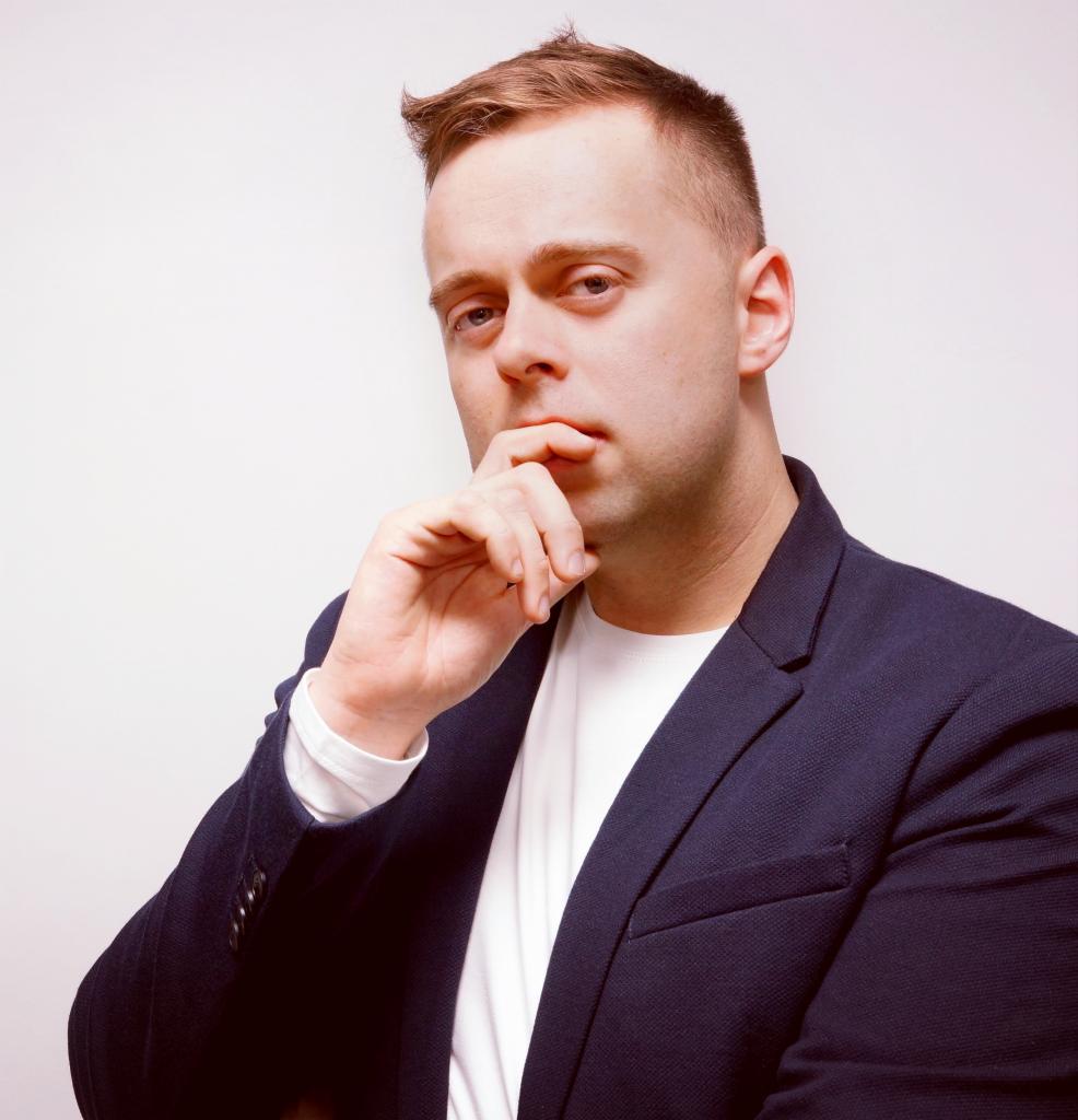 Portret mężczyzny w marynarce, zamyślony, z ręką przy ustach, na jasnym tle.