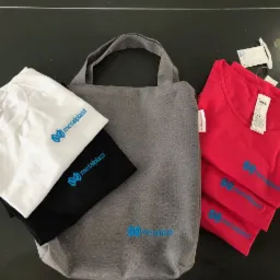 Zestaw firmowy: biała, czarna i trzy czerwone koszulki polo z niebieskim haftowanym logo 'metalplast' ułożone obok szarej torby materiałowej z tym samym haftem, na ciemnym tle.