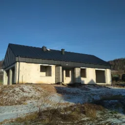 Surowy stan domu jednorodzinnego z białego bloczka i ciemnym dachem na wzniesieniu, w tle las i błękitne niebo.