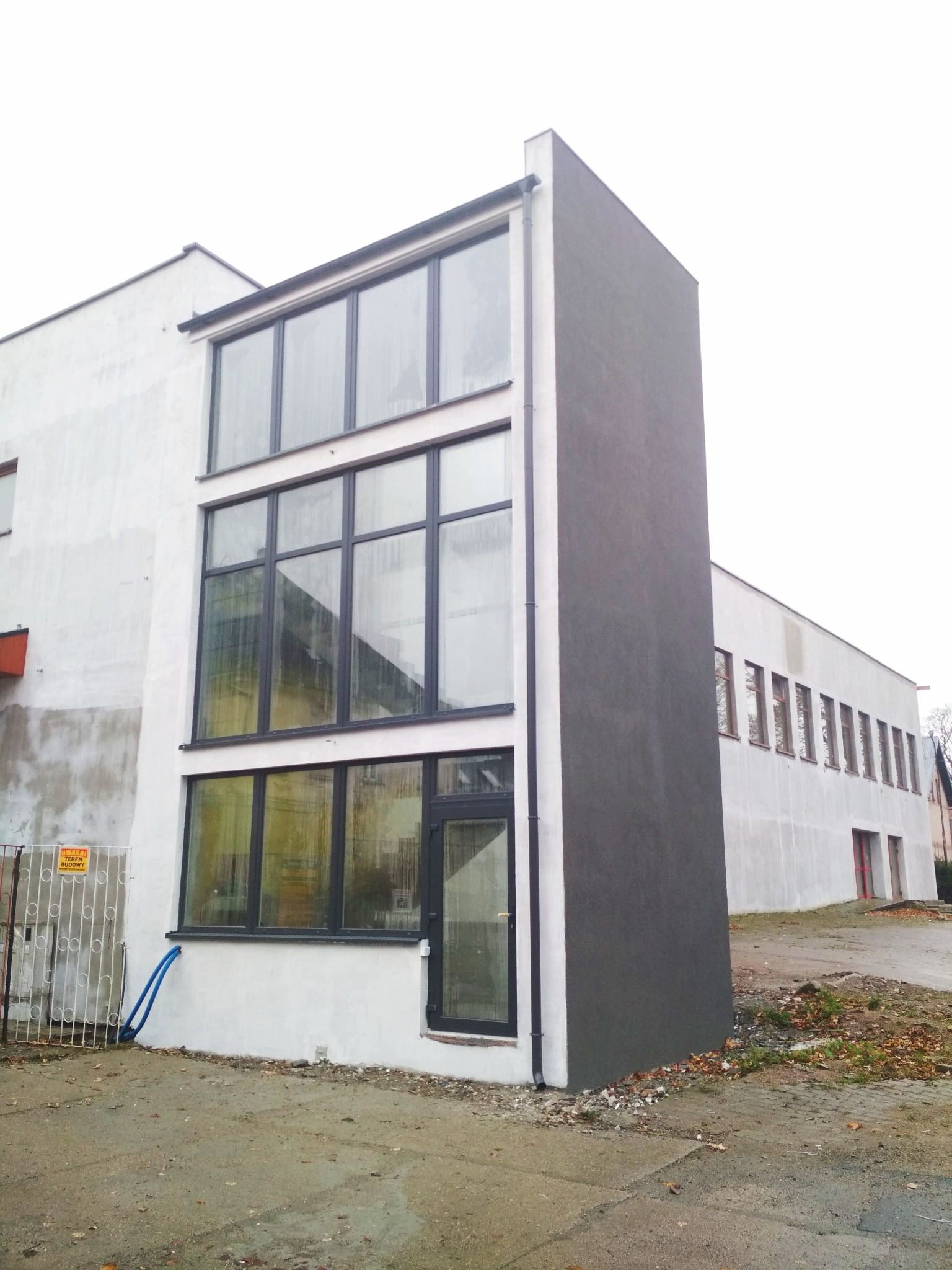 Trzykondygnacyjny budynek z dużymi oknami w czarnych ramach, część fasady w surowym, szarym tynku, widoczne rury na zewnątrz i tablica informacyjna o terenie budowy.