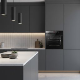Kors Meble - Nowoczesna, szara kuchnia z wyspą i wbudowanym piekarnikiem. Minimalistyczne oświetlenie podkreśla gładkie fronty szafek i biały blat. Ujęcie z bliska na detale.