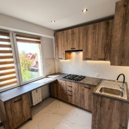 Kors Meble - Kompaktowa kuchnia z drewnianymi frontami i blatem w odcieniach szarości, z oknem i widokiem na zieleń. Uwagę zwraca nietypowe przedłużenie blatu pod oknem.