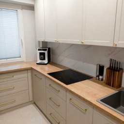 Kors Meble - Jasna kuchnia na wymiar z drewnianym blatem, złotymi uchwytami i płytą indukcyjną. Minimalistyczny design, widok na okno z żaluzjami.