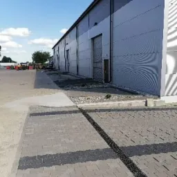 Szara kostka brukowa ułożona przed budynkiem przemysłowym z widocznym systemem odprowadzania wody w postaci czarnej kratki ściekowej, jasne niebo w tle.