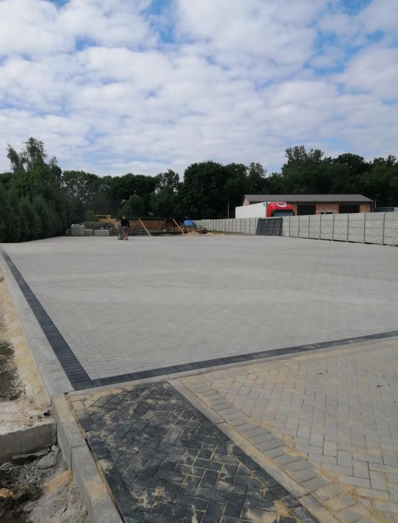 Rozległy plac wyłożony jasną kostką brukową z ciemnym obramowaniem, widoczny fragment starej nawierzchni, w tle zieleń i budynek magazynowy z czerwonym elementem dekoracyjnym.