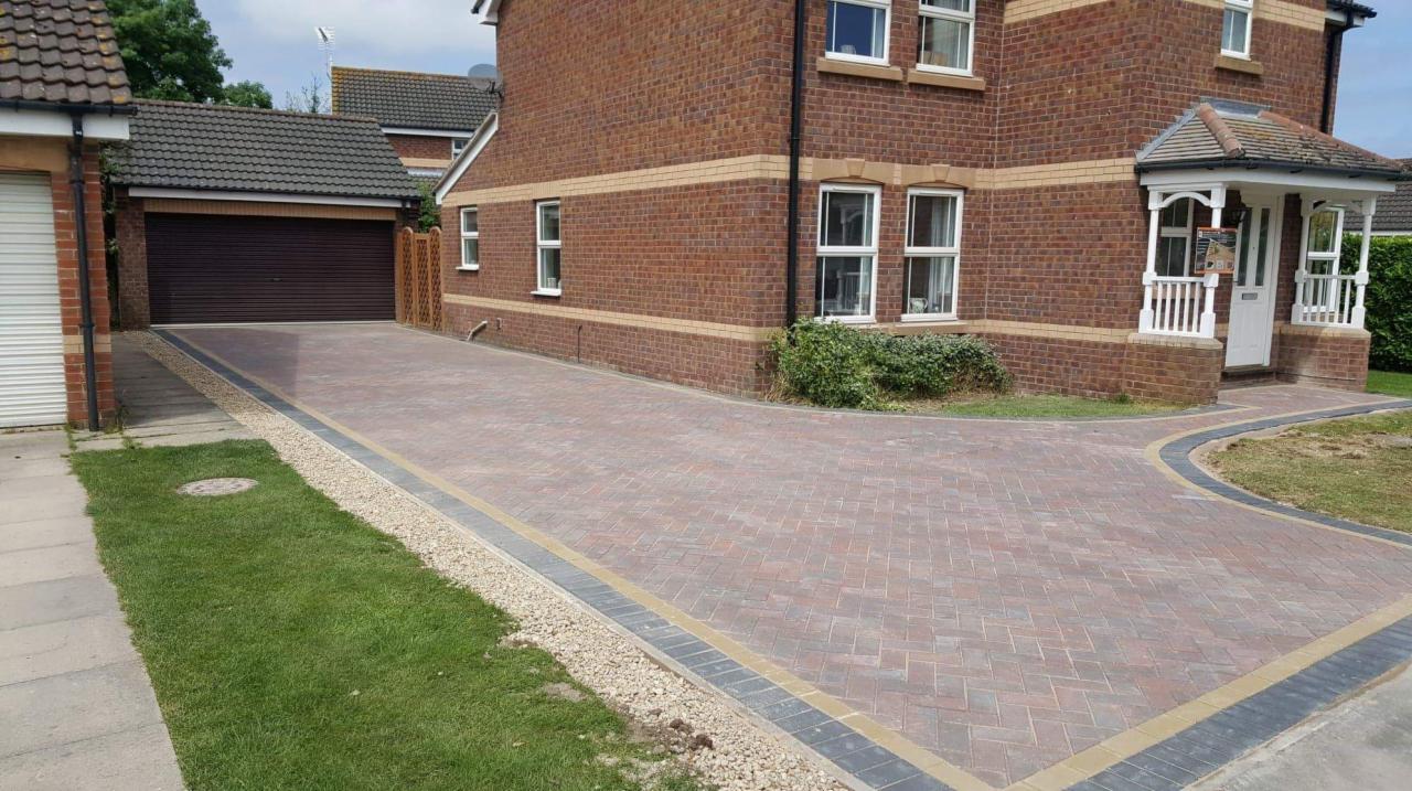 Nowa, wzorzysta nawierzchnia z czerwono-brązowej kostki brukowej prowadząca od garażu do wejścia domu z cegły, obrzeża wykończone drobnym żwirem.