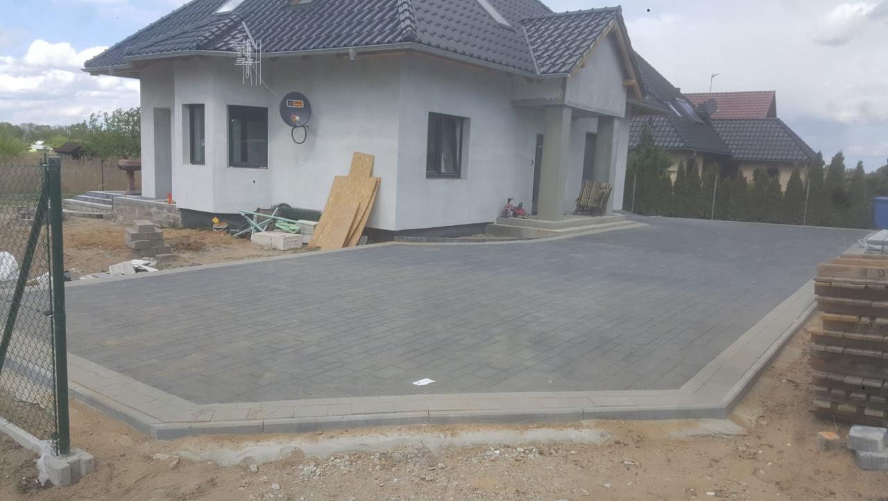 Ułożona szara kostka brukowa na podjeździe przed domem w trakcie budowy, widoczne deski, gruz i niezakończone prace wykończeniowe.