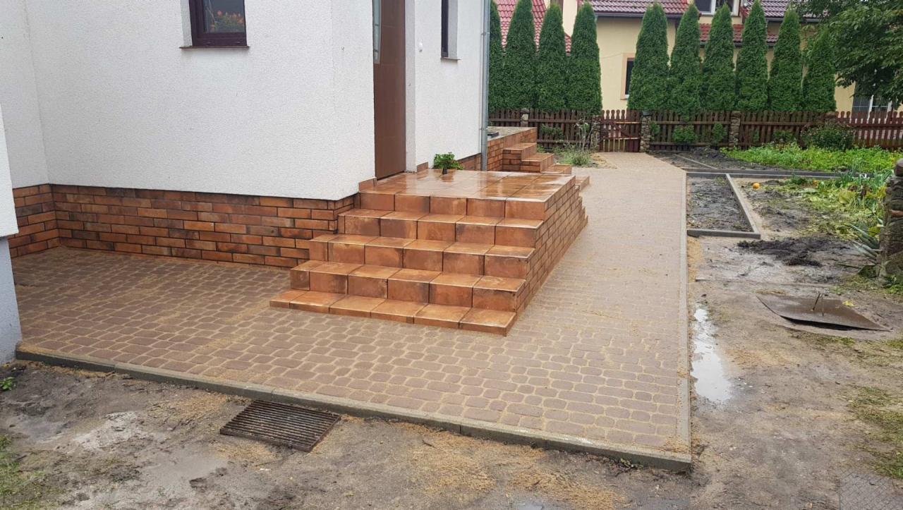 Nowo ułożona, mokra kostka brukowa w odcieniach brązu tworzy chodnik i schody wejściowe do domu z ceglaną podmurówką. Widoczny fragment ogrodu z zielonymi tujami w tle.